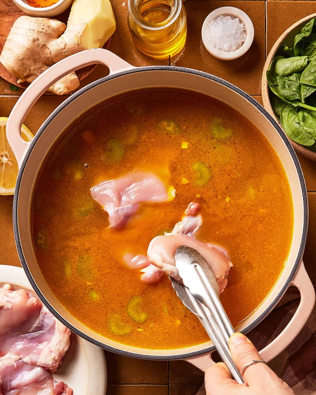 A hand uses tongs to add raw chicken pieces into a pot of broth with visible celery slices. Other ingredients like spinach, ginger, salt, and oil are arranged around the pot.