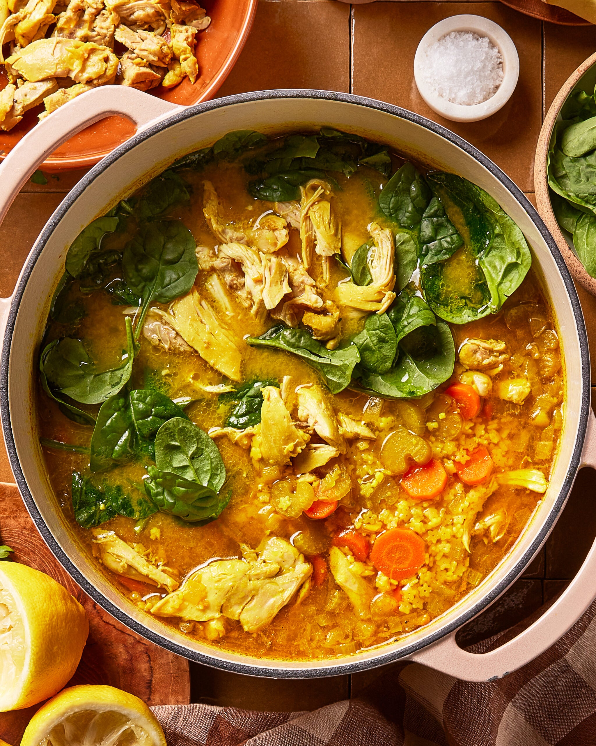 A pot of chicken soup with leafy spinach, carrots, and yellow broth sits on a table. Lemon and a bowl of salt are nearby, along with a plate of shredded chicken.
