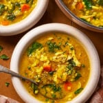A bowl of chicken and vegetable soup with rice, carrots, spinach, and herbs, shown with a spoon and another bowl partially visible nearby on a checkered cloth.