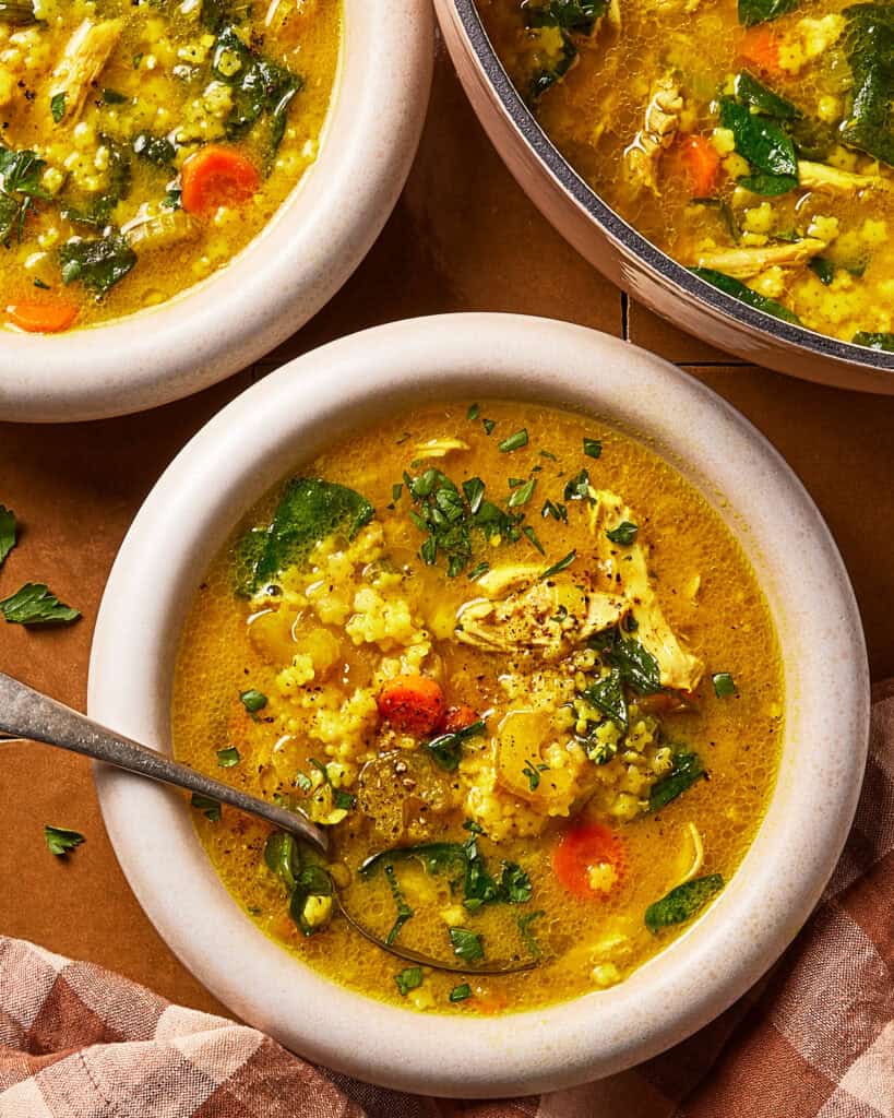 A bowl of chicken and vegetable soup with rice, carrots, spinach, and herbs, shown with a spoon and another bowl partially visible nearby on a checkered cloth.