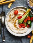 A bowl of the healthy white bean dip, topped with chopped cucumbers and tomatoes. A spoon can be seen resting next to the bowl.