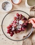 A clear bowl with the grapes inside of it, and a hand pouring the sea salt and baking soda on top of the grapes.