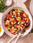 A large bowl of the ultimate fruit salad, sitting on a countertop, with serving spoons sitting inside of the bowl