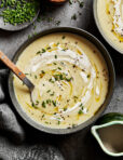 A bowl of the cozy potato leek soup, sitting on a countertop with a spoon sitting in the bowl