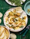 A bowl of the fried pickle dip, sitting on a countertop with a ruffled potato chip sitting in the bowl