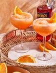 A glass of the easy frozen aperol spritz, sitting on a countertop topped with an orange slice and a bottle of aperol in the background