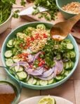 A vibrant salad in a light blue bowl features sliced cucumbers, red onion, chopped red chili peppers, cilantro, and peanuts. A spoon is held above, pouring dressing over the salad. Fresh herbs, a knife, and a partially cut lime are visible around the bowl.