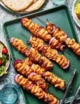 A green tray displays several skewers of grilled chicken and red onions. Tongs lie on the tray next to the skewers. In the background, there is a salad bowl with tomatoes and greens, a partially visible stack of pita bread, and glasses of water.