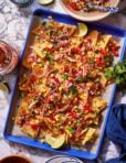 A colorful sheet pan of loaded nachos topped with shredded meat, melted cheese, diced tomatoes, corn kernels, sliced red onions, chopped cilantro, and lime wedges. The nachos sit on a parchment paper-lined blue tray set on a marble surface.