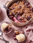 A round baking dish filled with cherry crisp topped with a crumbly, golden-brown streusel. A large serving spoon is placed inside. Two small bowls of cherry crisp topped with scoops of vanilla ice cream are beside it. Silver spoons and a pink cloth are nearby.