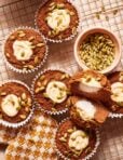 A cooling rack holds six pumpkin muffins, five of them whole and one cut in half to show the creamy filling. The muffins are topped with pumpkin seeds and are placed next to a small bowl filled with additional pumpkin seeds on a patterned napkin.