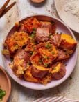 A bowl of crispy roasted sweet potatoes garnished with chopped green onions. The chunks are golden brown with a textured, crispy coating. They sit on a light background, creating an appetizing presentation.