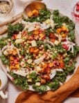 A vibrant salad with shredded chicken, kale, roasted butternut squash, chickpeas, apple cubes, and pomegranate seeds. It is served on a white plate with wooden utensils and a brown napkin beside it.