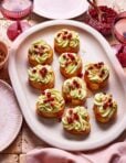 A platter of eight crostini topped with swirls of green cream and pomegranate seeds, surrounded by glasses of rosé and bowls of pomegranate seeds and chopped nuts. The setting is on a tiled surface with pink napkins.
