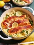 A white pot filled with cooked salmon filets on a bed of pearled couscous, spinach, and sliced lemons. A smaller bowl with couscous is in the background, alongside whole lemons and a small dish of pepper. A yellow cloth is nearby.