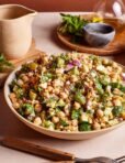 A vibrant and fresh salad in a bowl featuring chickpeas, diced cucumbers, red onions, fresh herbs, and crumbled feta cheese. A jug and fresh herbs are visible in the background, giving a homely kitchen vibe.