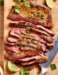 Sliced grilled beef steak garnished with chopped herbs and jalapeños on a wooden cutting board, accompanied by lime wedges and cilantro sprigs. A knife lies beside the steak.