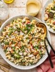 A large bowl of pasta salad with grilled chicken, radish slices, chickpeas, green peas, asparagus, herbs, and feta cheese. Two serving spoons rest in the bowl; a glass of dressing and a pink napkin are nearby.