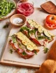 Two open-faced sandwiches with turkey, zucchini slices, fresh arugula, pickled onions, and tomato on flatbread, served on a wooden board with a small bowl of salt and pepper, arugula, and a glass of pickled onions nearby.