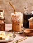 A glass of iced coffee with a gold metal straw sits on a woven coaster, surrounded by jars of coffee, a pitcher, and a plate of sliced bananas on a beige surface.