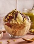 A glass bowl of pistachio ice cream topped with chocolate sauce, chopped pistachios, and shredded pastry, with a jar of pistachio spread and a spoon in the background.