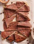 Sliced chocolate-covered bars on parchment paper, sprinkled with flaky sea salt, with two knives and bowls of salt and chocolate chips nearby.