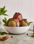 Two scoops of green ice cream in a white bowl, topped with chocolate sauce and cookie crumbles, garnished with mint leaves. A vase with mint and a stack of cookies are in the background.