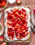 A rectangular sheet cake topped with whipped cream and fresh sliced strawberries sits on a pink tile surface. Plates, forks, a knife, and bowls of strawberries surround the cake.