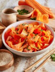 A bowl of carrot ribbons garnished with sesame seeds and chopped herbs sits on a table, surrounded by fresh carrots, herbs, a small pitcher, and a bowl of sesame seeds. Chopsticks lie beside the bowl.
