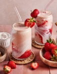 Two glasses of strawberry and cream smoothie with swirled red and white layers, topped with fresh strawberries and clear straws. Fresh strawberries and a bowl of cream are nearby on a pink tiled surface.