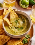A bowl of green salsa garnished with cilantro is surrounded by tortilla chips. Glasses of beer, a green tomato, and a jalapeño are in the background on a wooden plate.