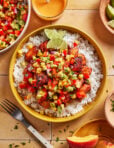 A bowl of white rice topped with grilled fish and colorful mango salsa, garnished with lime wedges. Surrounding the bowl are a fork, extra salsa, sauce, lime slices, and herbs on a tiled surface.