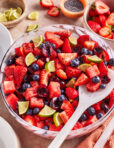 A large bowl of fresh fruit salad with strawberries, blueberries, watermelon, cherries, and lime wedges. A white serving spoon rests in the bowl, with extra fruit and lime slices on the table beside it.