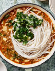 A pot of noodle soup with uncooked rice noodles, leafy greens, minced meat, and chopped vegetables in a rich, reddish broth, placed on a green textured surface.
