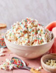 A bowl of popcorn topped with colorful sprinkles sits on a pink surface. A spoon with more sprinkles, a jar of sprinkles, white chocolate chips, and a peach napkin are nearby.
