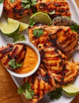 Grilled chicken thighs served on a white platter with lime wedges, fresh cilantro, and a small bowl of orange dipping sauce. The chicken has visible grill marks and a golden, slightly charred appearance.