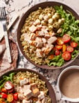 A Copycat Sweetgreen Chicken Caprese Bowl filled with quinoa, grilled chicken, cherry tomatoes, arugula, mozzarella balls, and creamy dressing sit on a marble surface next to a napkin, forks, and a bowl of extra dressing.