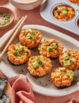 A plate of six Spicy California Roll Baked Sushi Cups topped with avocado and chopped chives, surrounded by chopsticks, a bowl of dipping sauce, a baking tray with more sushi cups, and a small dish of chives.