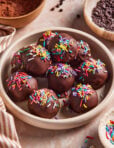 A bowl filled with chocolate-coated balls topped with colorful sprinkles sits on a marble surface, surrounded by bowls of chocolate chips, cocoa powder, and a striped cloth. (Viral High Protein Brownie Bites)