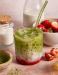 A glass of iced matcha latte with layers of green matcha, milk, and red strawberry puree sits on a table with a glass straw. Surrounding it are bowls of sugar, strawberries, a bottle of milk, and a dish of matcha powder.