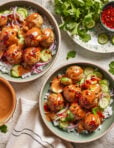 Two bowls of rice topped with chicken dumpling meatballs, sliced cucumbers, red onion, and creamy sauce, garnished with chili flakes. Fresh cilantro, cucumber slices, and a bowl of sauce are arranged nearby.
